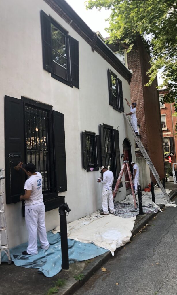 Commercial painters applying fresh white paint to a historic building's exterior, with ladders and drop cloths visible, showcasing professional painting services in a residential area.