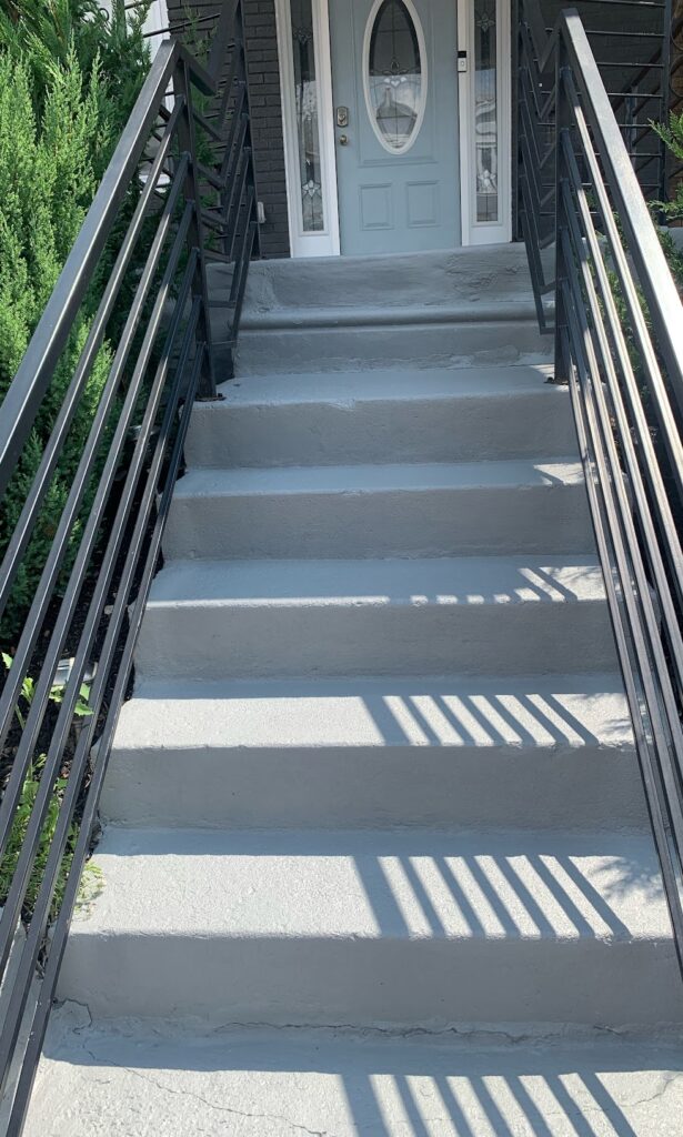Staircase leading to a blue front door, featuring freshly painted gray steps and black railings, surrounded by greenery, emphasizing quality and craftsmanship in commercial painting services.